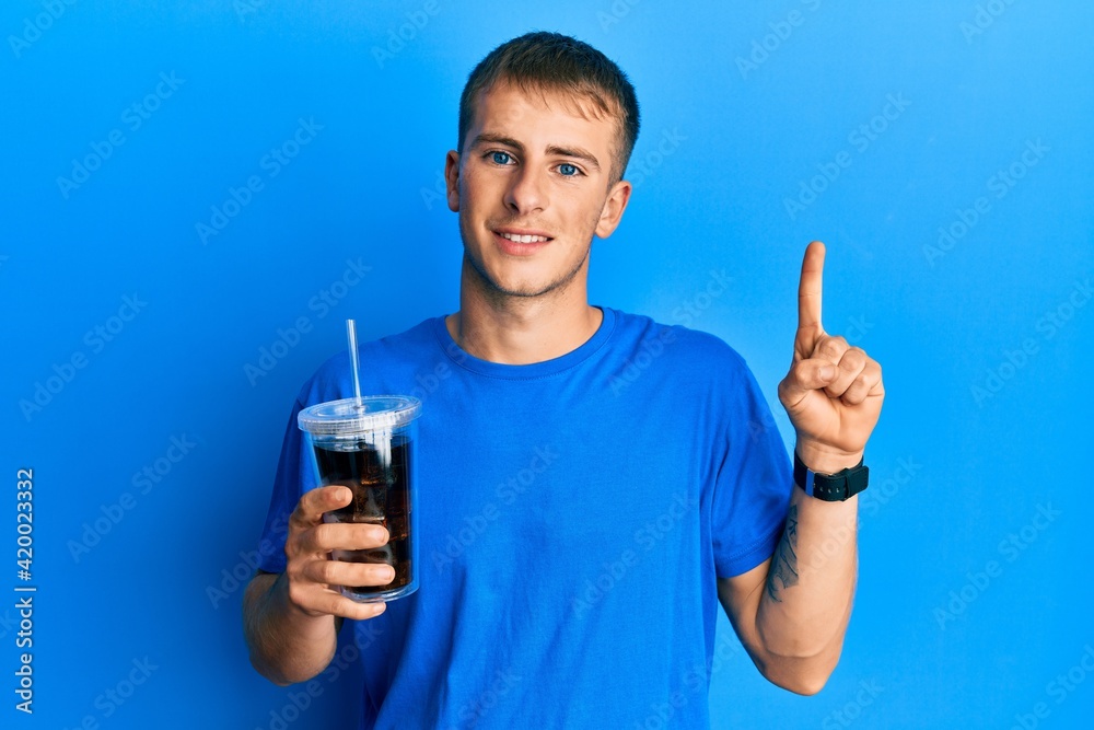 Young caucasian man drinking glass of soda beverage smiling with an idea or question pointing finger with happy face, number one Young caucasian man drinking glass of soda beverage smiling with an idea or question pointing finger with happy face, number one