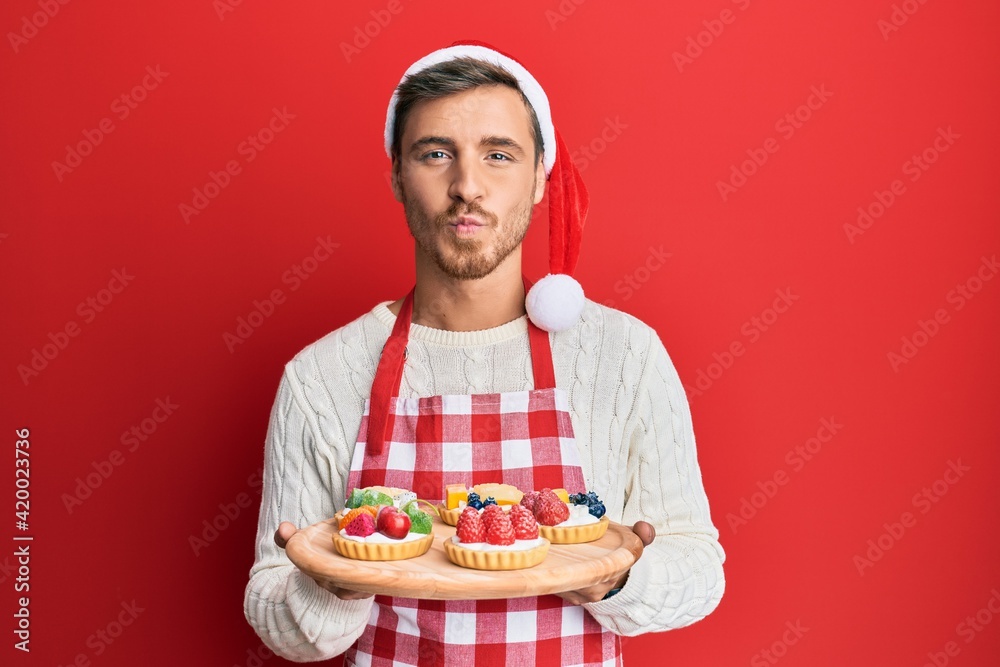 Handsome caucasian man wearing baker apron and christmas hat holding cakes looking at the camera blowing a kiss being lovely and sexy. love expression. Handsome caucasian man wearing baker apron and christmas hat holding cakes looking at the camera blowing a kiss being lovely and sexy. love expression.