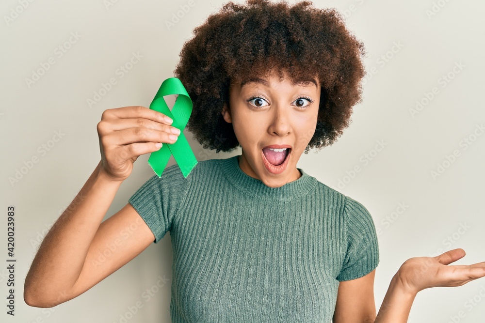 Young hispanic girl holding support green ribbon celebrating victory with happy smile and winner expression with raised hands Young hispanic girl holding support green ribbon celebrating victory with happy smile and winner expression with raised hands
