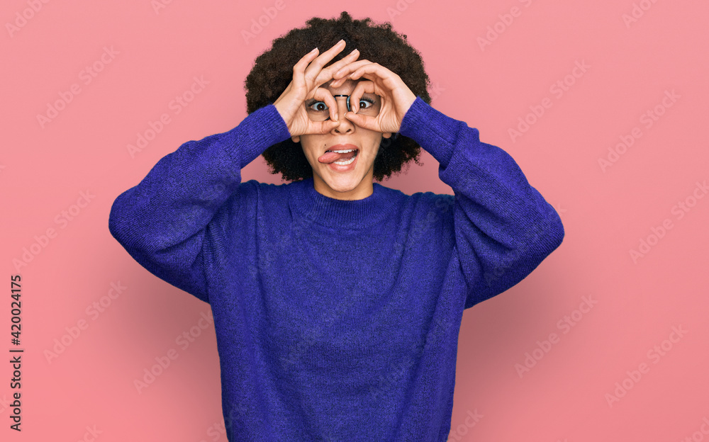 Young hispanic girl wearing casual winter sweater and glasses doing ok gesture like binoculars sticking tongue out, eyes looking through fingers. crazy expression.