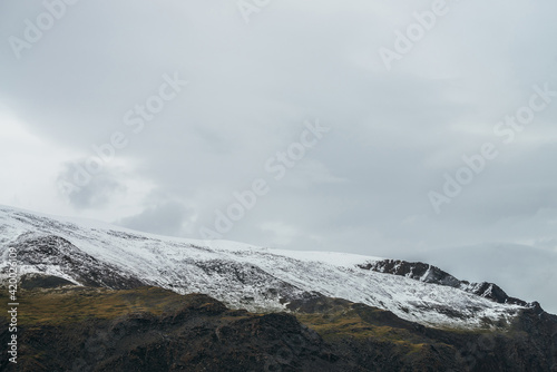 Minimalist highlands landscape with snow-capped mountains under cloudy sky. Snowbound high mountain range in overcast weather. Atmospheric minimalism with green mountains, white snow and gray sky.