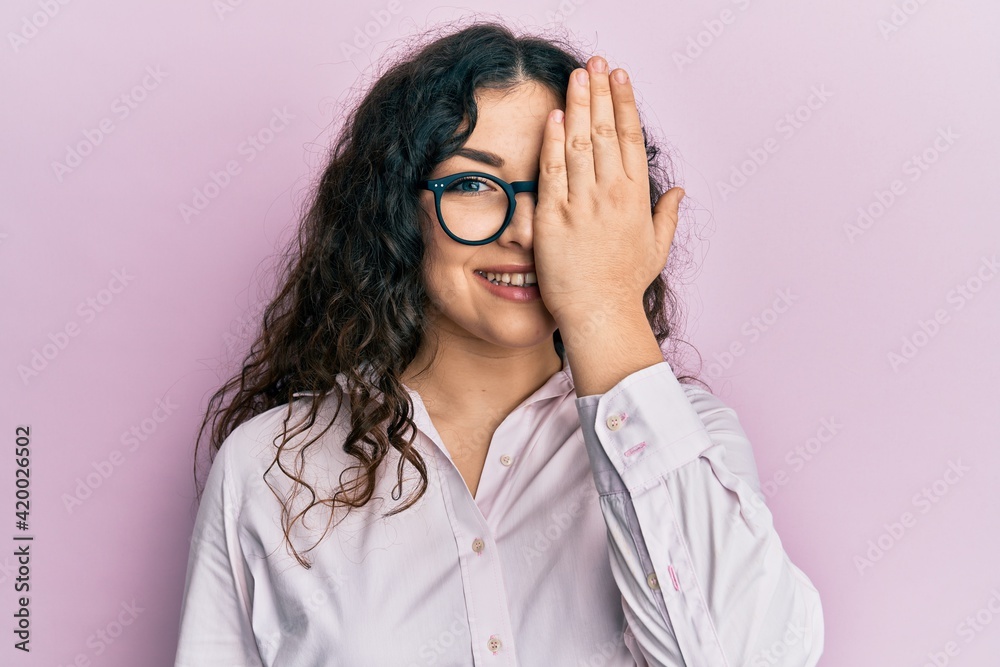 Young brunette woman with curly hair wearing casual clothes and glasses covering one eye with hand, confident smile on face and surprise emotion. Young brunette woman with curly hair wearing casual clothes and glasses covering one eye with hand, confident smile on face and surprise emotion.