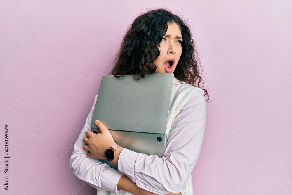 Fototapeta premium Young brunette woman with curly hair hugging laptop with love angry and mad screaming frustrated and furious, shouting with anger. rage and aggressive concept.