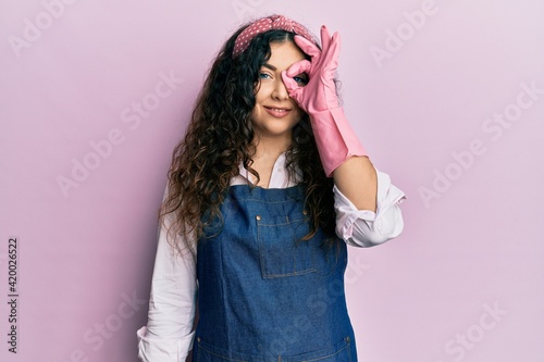 Young brunette woman with curly hair wearing cleaner apron and gloves doing ok gesture with hand smiling, eye looking through fingers with happy face.