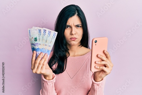 Young hispanic woman using smartphone holding colombian pesos banknotes skeptic and nervous, frowning upset because of problem. negative person.
