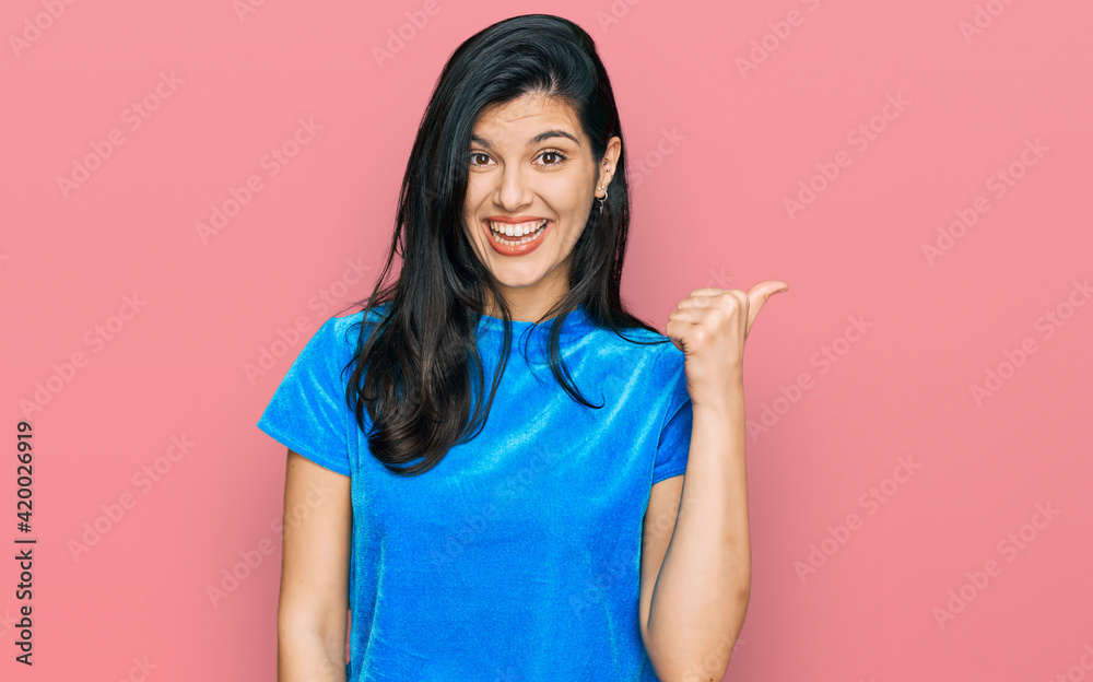 Young hispanic woman wearing casual clothes smiling with happy face looking and pointing to the side with thumb up. Young hispanic woman wearing casual clothes smiling with happy face looking and pointing to the side with thumb up.
