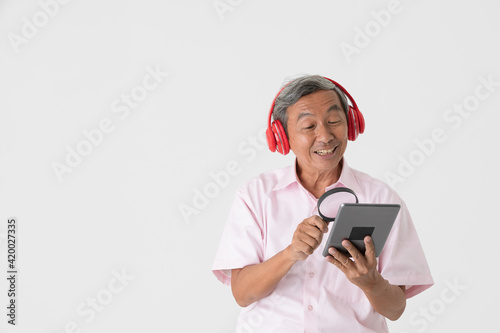 Portrait of a good-looking active senior older Asian man holding and wearing red headphone and listening to music while using a magnifying glass watch tablet computer screen with joyfully and happy © Bangkok Click Studio