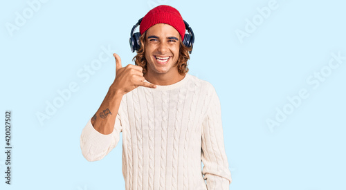 Young hispanic man listening to music using headphones smiling doing phone gesture with hand and fingers like talking on the telephone. communicating concepts.