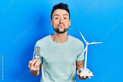 Young hispanic man holding windmill and led lightbulb looking at the camera blowing a kiss being lovely and sexy. love expression.