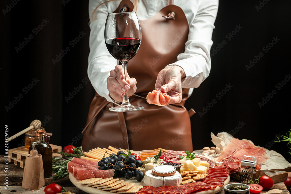 Hands holding a glass of wine and a wooden board with different kinds of cheese and ham, prosciutto, jamon salami, Antipasto