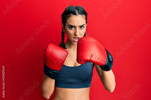 Young brunette girl using boxing gloves in shock face, looking skeptical and sarcastic, surprised with open mouth