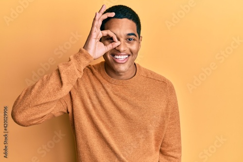 Young handsome hispanic man wearing casual winter sweater doing ok gesture with hand smiling, eye looking through fingers with happy face. © Krakenimages.com