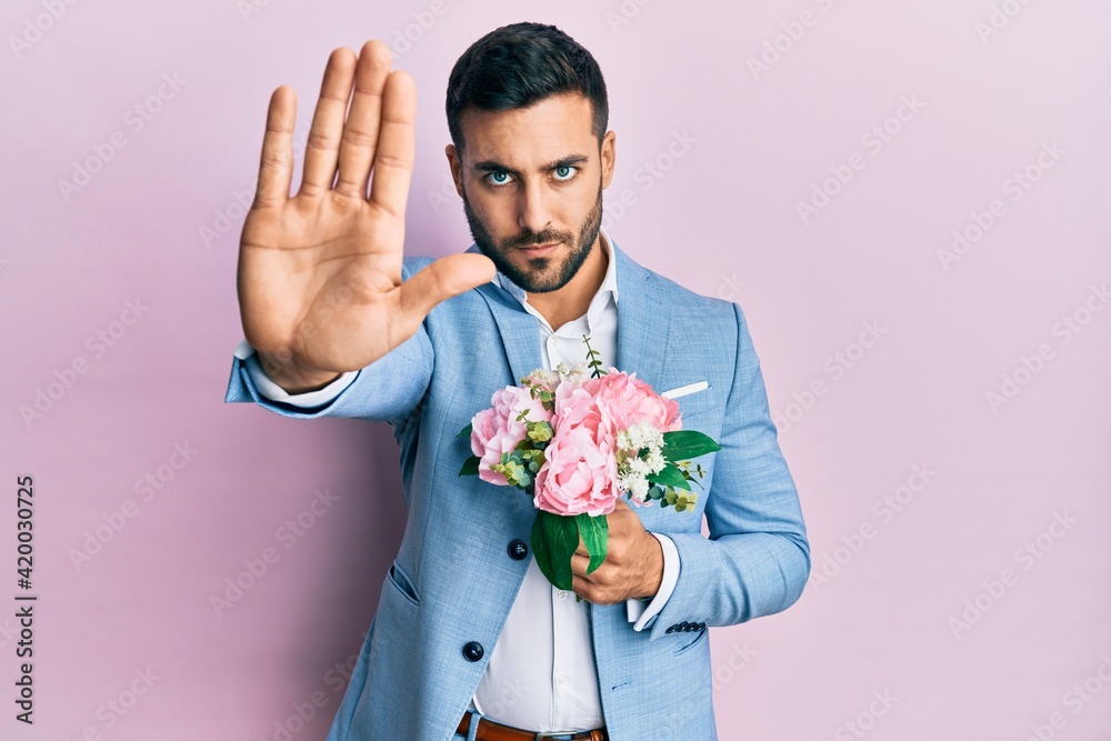 Young hispanic businessman wearing business jacket holding flowers with open hand doing stop sign with serious and confident expression, defense gesture