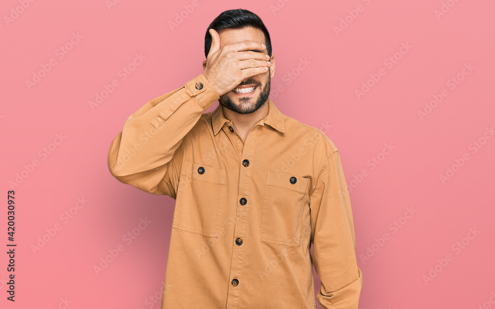 Young hispanic man wearing casual clothes smiling and laughing with hand on face covering eyes for surprise. blind concept.