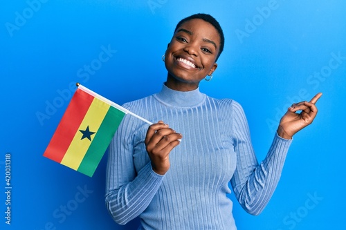 Young african american woman holding ghana flag smiling happy pointing with hand and finger to the side