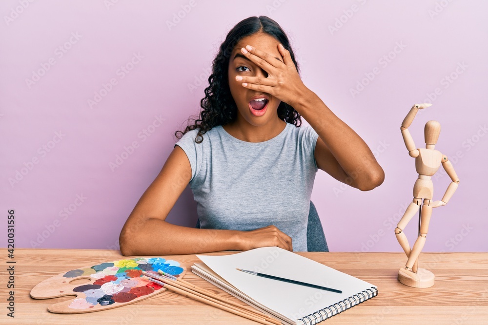 Young african american girl artist sitting at studio table peeking in shock covering face and eyes with hand, looking through fingers afraid Young african american girl artist sitting at studio table peeking in shock covering face and eyes with hand, looking through fingers afraid