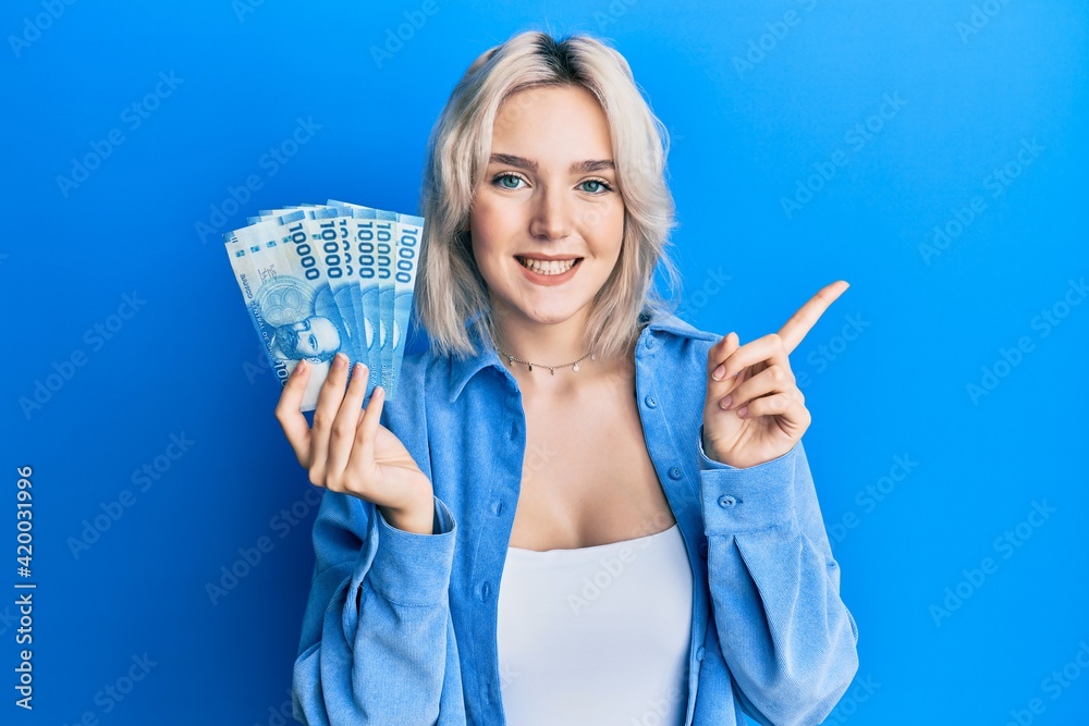 Young blonde girl holding 1000 chilean pesos smiling happy pointing with hand and finger to the side Young blonde girl holding 1000 chilean pesos smiling happy pointing with hand and finger to the side