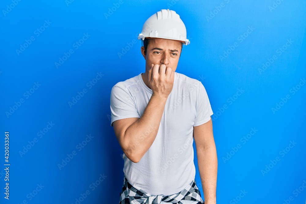 Handsome young man wearing builder uniform and hardhat looking stressed and nervous with hands on mouth biting nails. anxiety problem.