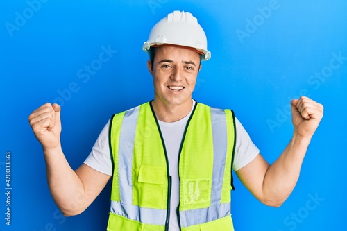 Handsome young man wearing safety helmet and reflective jacket screaming proud, celebrating victory and success very excited with raised arms