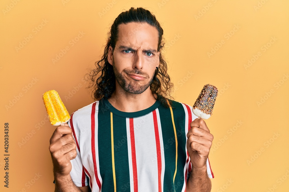 Young handsome man with long hair eating two ice cream skeptic and nervous, frowning upset because of problem. negative person. Young handsome man with long hair eating two ice cream skeptic and nervous, frowning upset because of problem. negative person.