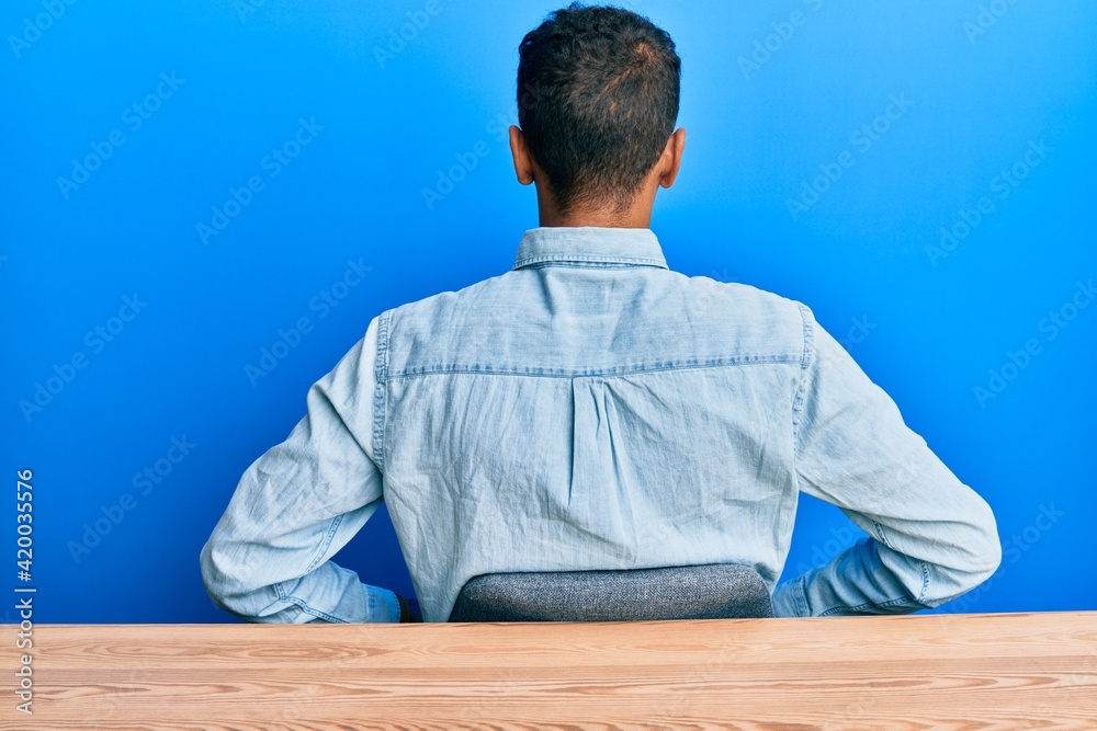 Fototapeta premium Young handsome african american man wearing casual clothes sitting on the table standing backwards looking away with arms on body