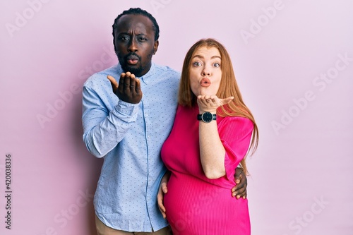 Young interracial couple expecting a baby, touching pregnant belly looking at the camera blowing a kiss with hand on air being lovely and sexy. love expression.