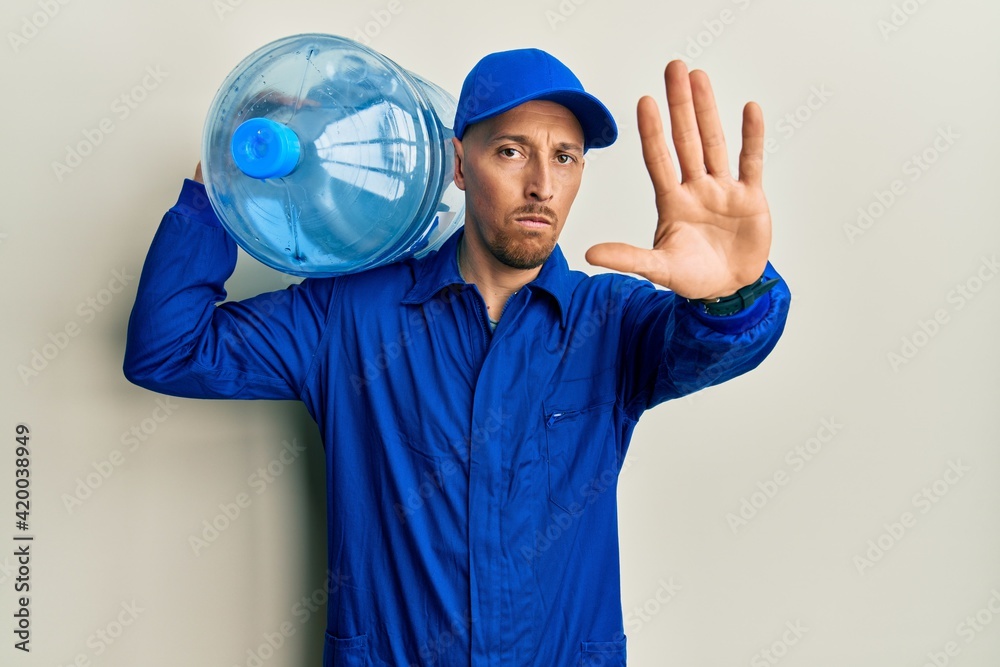 Bald courier man with beard holding a gallon bottle of water for delivery with open hand doing stop sign with serious and confident expression, defense gesture
