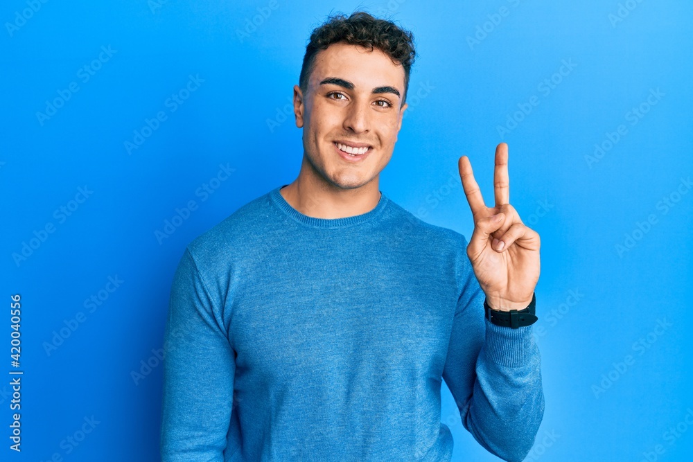 Hispanic young man wearing casual winter sweater smiling looking to the camera showing fingers doing victory sign. number two. Hispanic young man wearing casual winter sweater smiling looking to the camera showing fingers doing victory sign. number two.