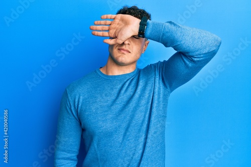 Hispanic young man wearing casual winter sweater covering eyes with arm, looking serious and sad. sightless, hiding and rejection concept