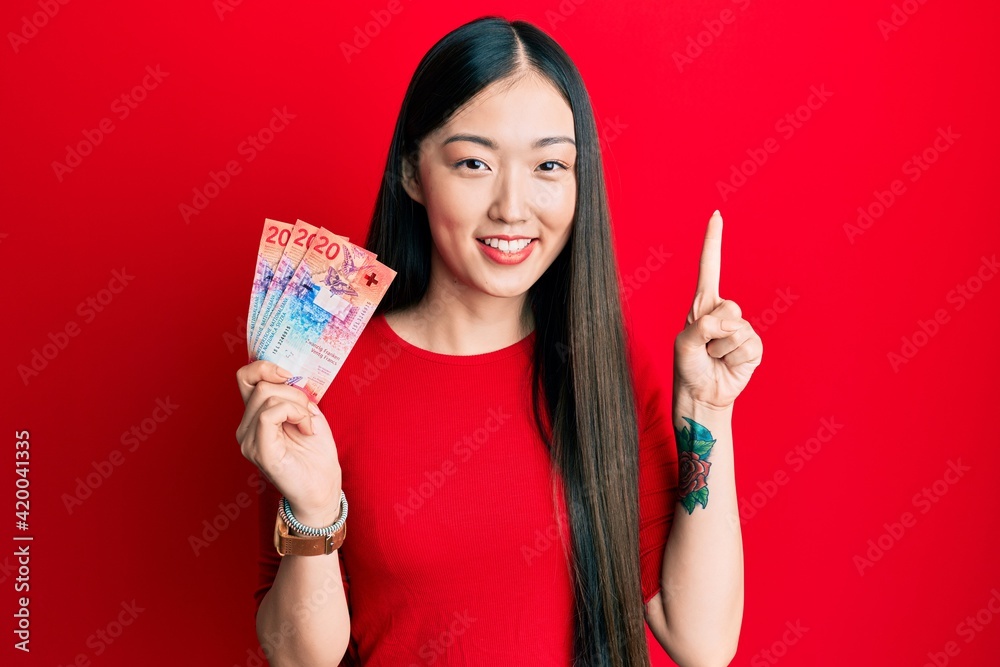 Young chinese woman holding 20 swiss franc banknotes smiling with an idea or question pointing finger with happy face, number one Young chinese woman holding 20 swiss franc banknotes smiling with an idea or question pointing finger with happy face, number one