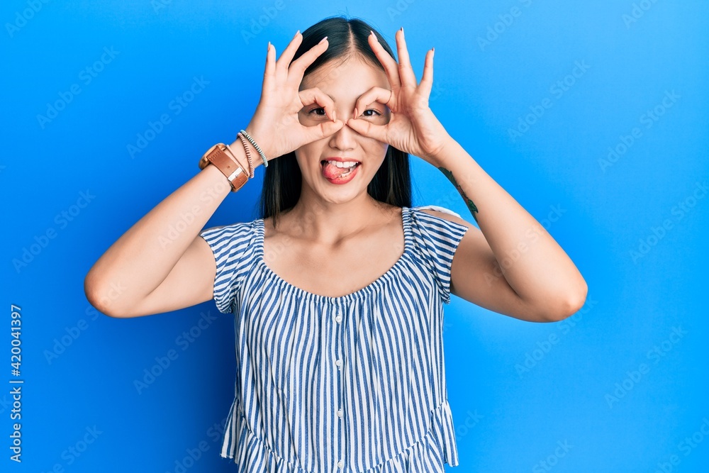 Young chinese woman wearing casual striped t-shirt doing ok gesture like binoculars sticking tongue out, eyes looking through fingers. crazy expression. Young chinese woman wearing casual striped t-shirt doing ok gesture like binoculars sticking tongue out, eyes looking through fingers. crazy expression.