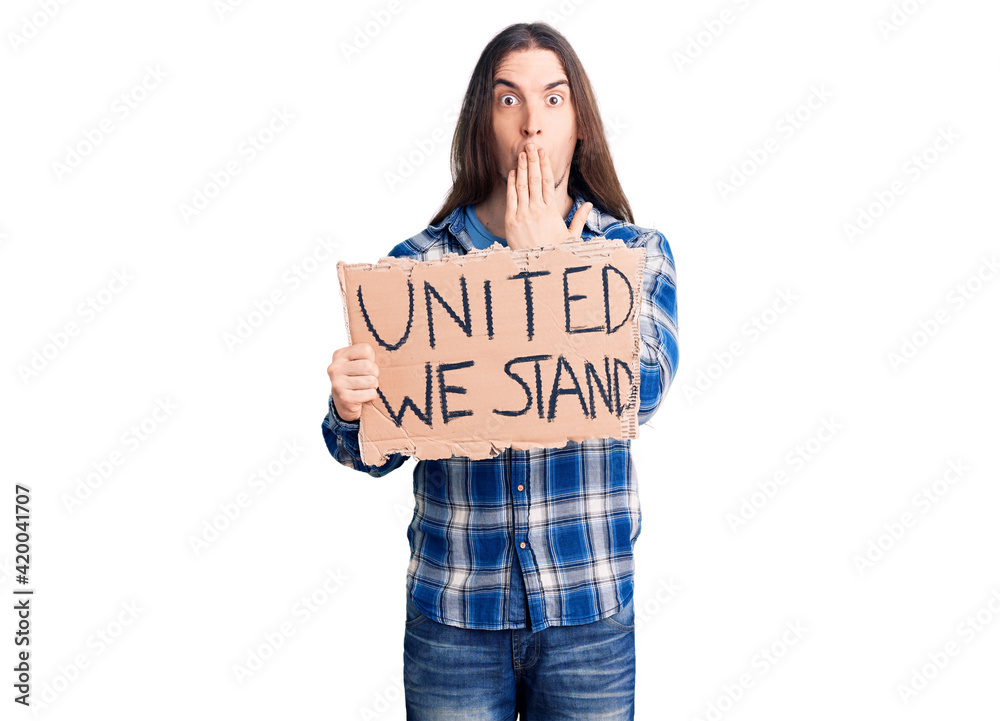 Young adult man with long hair holding united we stand banner covering mouth with hand, shocked and afraid for mistake. surprised expression Young adult man with long hair holding united we stand banner covering mouth with hand, shocked and afraid for mistake. surprised expression