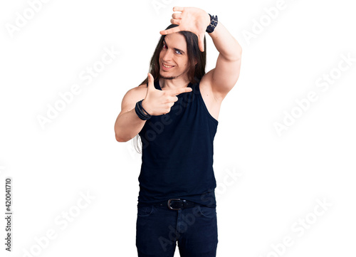 Young adult man with long hair wearing rocker style with black clothes and contact lenses smiling making frame with hands and fingers with happy face. creativity and photography concept.