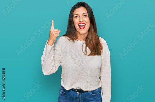 Beautiful hispanic woman wearing casual sweater pointing finger up with successful idea. exited and happy. number one.