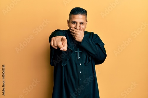 Young latin priest man standing over yellow background laughing at you  pointing finger to the camera with hand over mouth  shame expression
