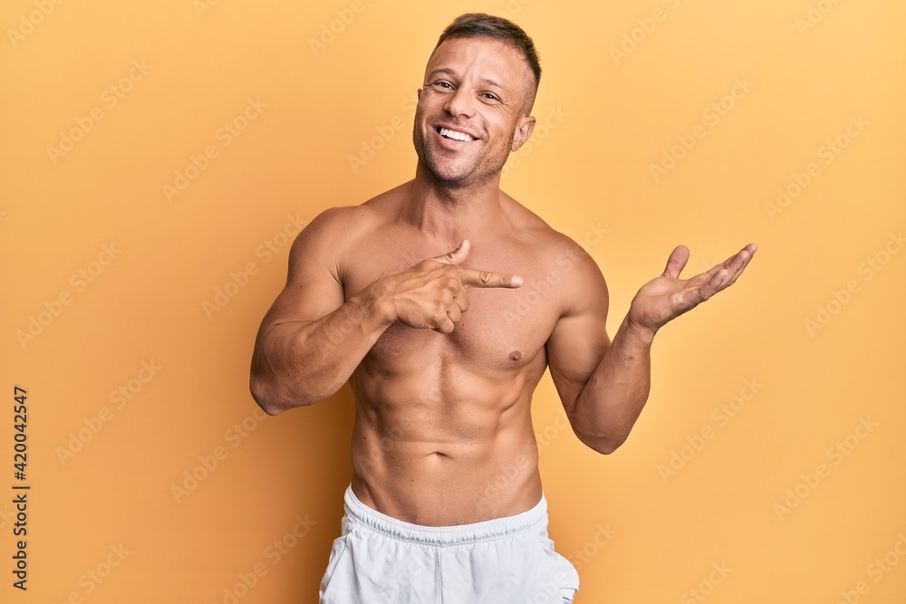 Handsome muscle man standing shirtless amazed and smiling to the camera while presenting with hand and pointing with finger.