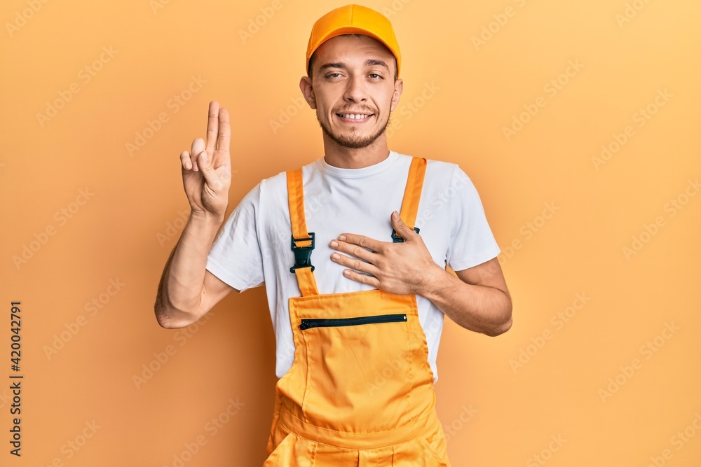 Hispanic young man wearing handyman uniform smiling swearing with hand on chest and fingers up, making a loyalty promise oath