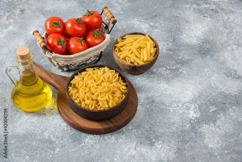 Wooden bowls of raw various pasta and fresh tomatoes on marble background