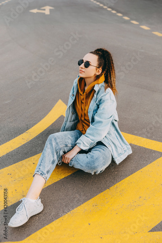 Young woman wearing denim clothes, sitting on an asphalted road.