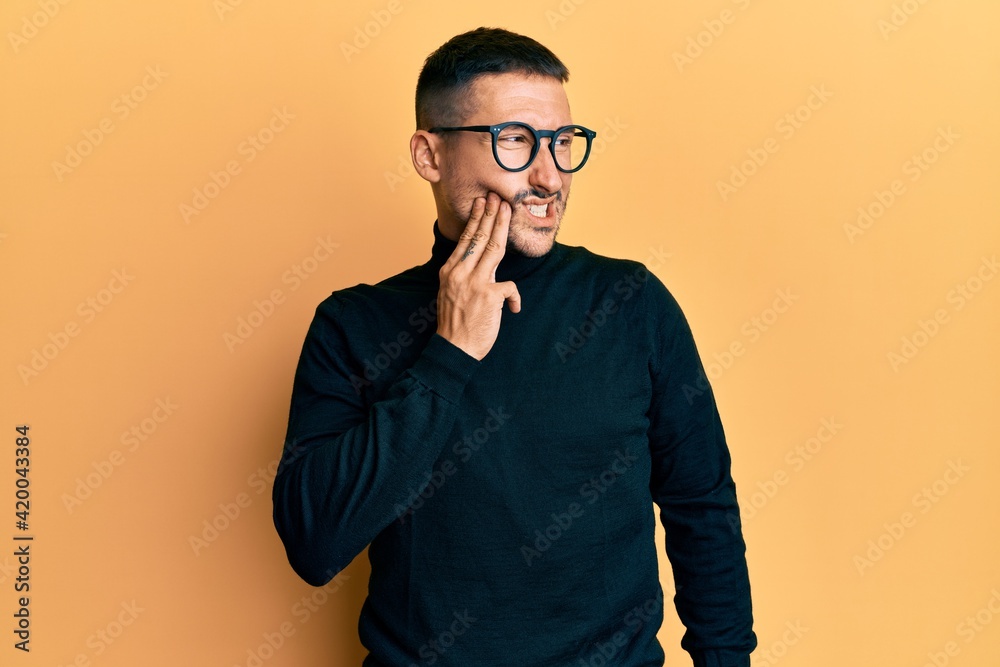 Fototapeta premium Handsome man with tattoos wearing turtleneck sweater and glasses touching mouth with hand with painful expression because of toothache or dental illness on teeth. dentist