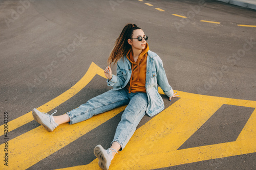 Young woman wearing denim clothes and sunglasses, sitting on an asphalted city road.