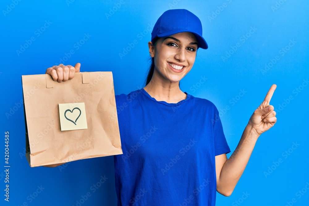 Young hispanic woman holding take away paper bag with heart reminder smiling happy pointing with hand and finger to the side Young hispanic woman holding take away paper bag with heart reminder smiling happy pointing with hand and finger to the side