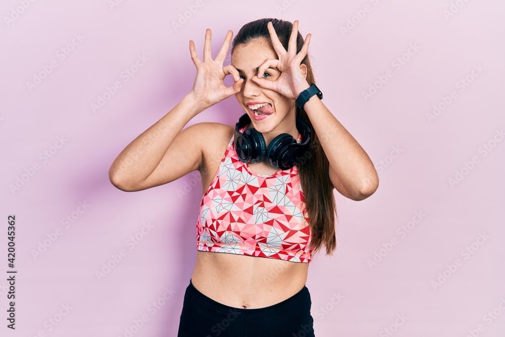 Young brunette teenager wearing gym clothes and using headphones doing ok gesture like binoculars sticking tongue out, eyes looking through fingers. crazy expression. Young brunette teenager wearing gym clothes and using headphones doing ok gesture like binoculars sticking tongue out, eyes looking through fingers. crazy expression.