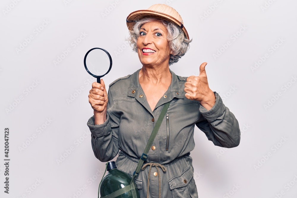 Senior grey-haired woman wearing explorer hat holding magnifying glass smiling happy and positive, thumb up doing excellent and approval sign Senior grey-haired woman wearing explorer hat holding magnifying glass smiling happy and positive, thumb up doing excellent and approval sign