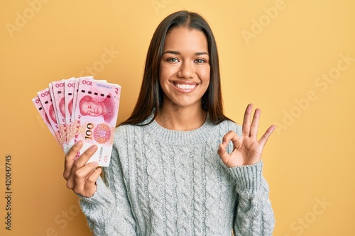 Beautiful hispanic woman holding 100 yuan chinese banknotes doing ok sign with fingers, smiling friendly gesturing excellent symbol