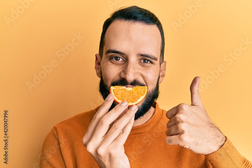 Young man with beard holding orange slice on mouth as funny smile smiling happy and positive  thumb up doing excellent and approval sign