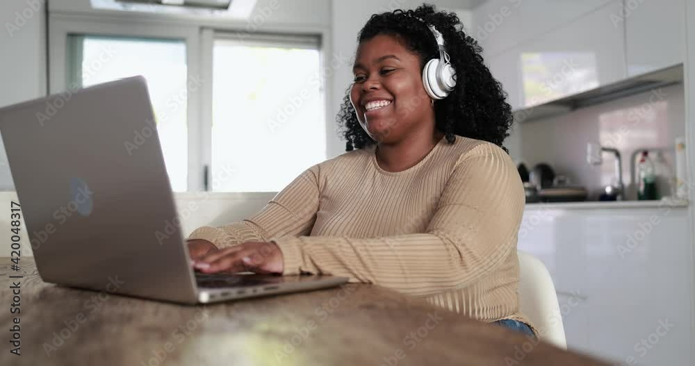 Happy african girl sitting inside kitchen at home using laptop computer and headphones 