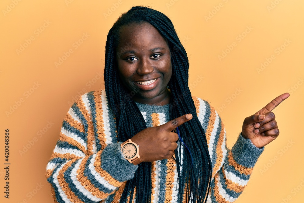Young black woman with braids wearing casual winter sweater smiling and looking at the camera pointing with two hands and fingers to the side. Young black woman with braids wearing casual winter sweater smiling and looking at the camera pointing with two hands and fingers to the side.