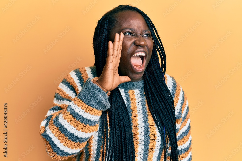 Young black woman with braids wearing casual winter sweater shouting and screaming loud to side with hand on mouth. communication concept. Young black woman with braids wearing casual winter sweater shouting and screaming loud to side with hand on mouth. communication concept.