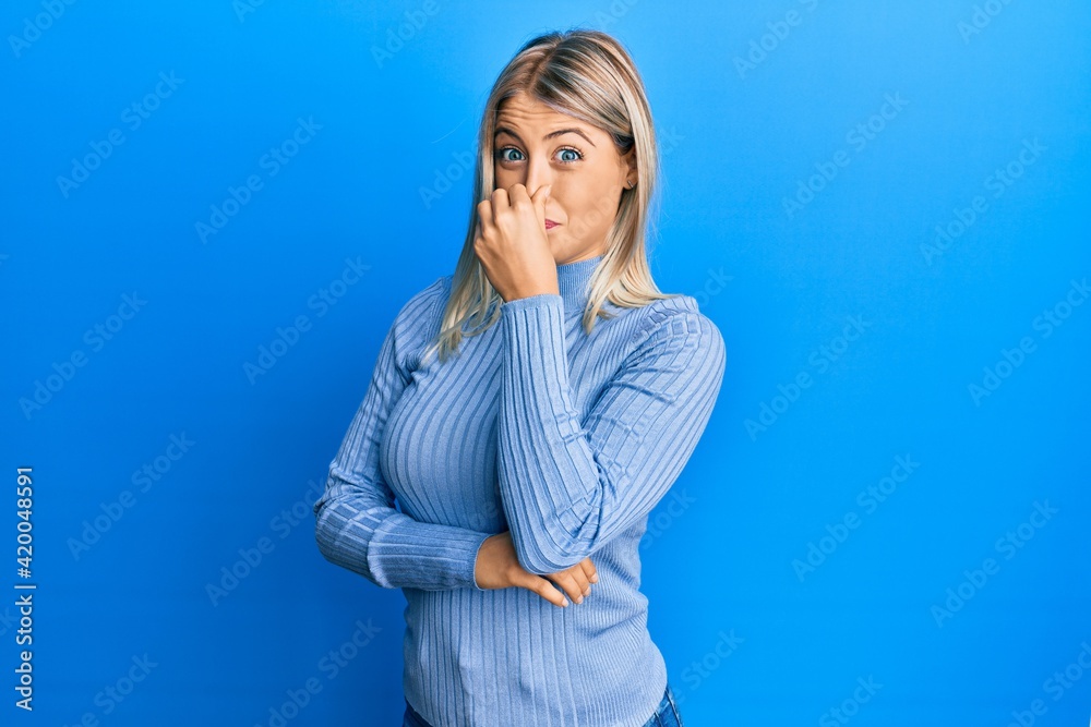 Beautiful blonde woman wearing casual clothes smelling something stinky and disgusting, intolerable smell, holding breath with fingers on nose. bad smell Beautiful blonde woman wearing casual clothes smelling something stinky and disgusting, intolerable smell, holding breath with fingers on nose. bad smell